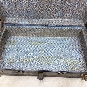 May include: An open, vintage trunk with a dark exterior and a patterned interior. The interior lining features a blue, yellow, and white geometric design. The trunk has metal hardware, including latches and hinges.