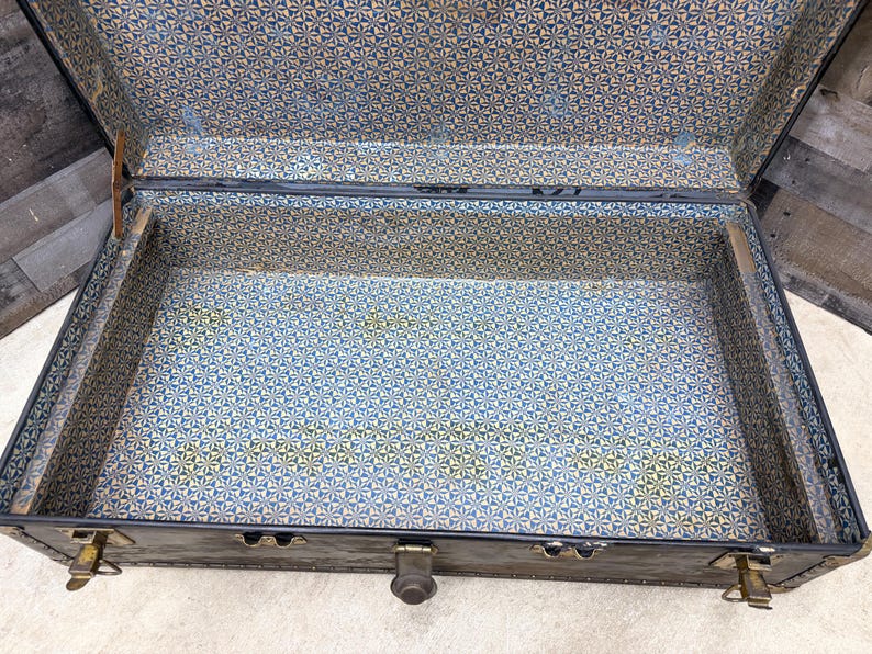 May include: An open, vintage trunk with a dark exterior and a patterned interior. The interior lining features a blue, yellow, and white geometric design. The trunk has metal hardware, including latches and hinges.