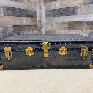 May include: A vintage black trunk with brass hardware. The trunk has a rectangular shape with rounded corners and is adorned with brass locks, hinges, and decorative studs. The trunk's exterior shows signs of wear, adding to its antique charm.