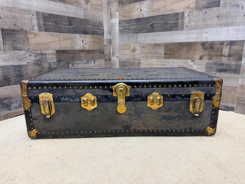 May include: A vintage black trunk with brass hardware. The trunk has a rectangular shape with rounded corners and is adorned with brass locks, hinges, and decorative studs. The trunk's exterior shows signs of wear, adding to its antique charm.