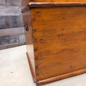 Op de afbeelding: Een houten kist met een gebogen bovenkant en metalen handvat. De kist heeft een warme bruine kleur met zichtbare houtnerf en zwaluwstaartverbindingen. De kist lijkt vintage of antiek te zijn.