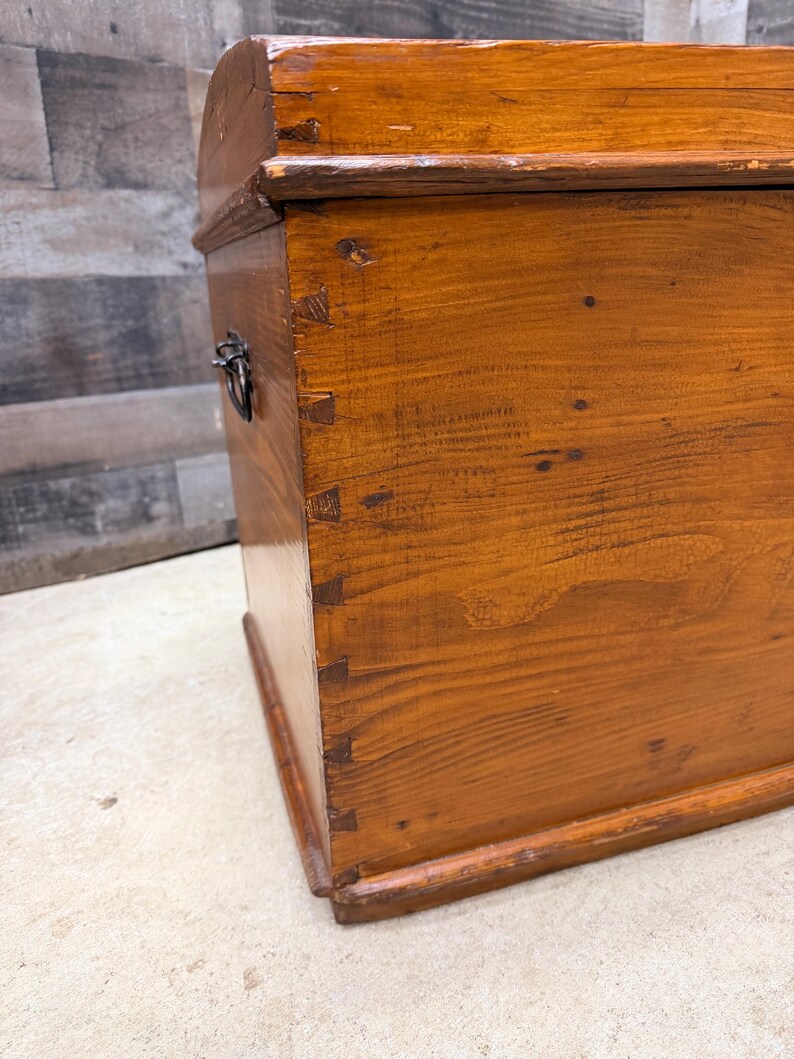 Op de afbeelding: Een houten kist met een gebogen bovenkant en metalen handvat. De kist heeft een warme bruine kleur met zichtbare houtnerf en zwaluwstaartverbindingen. De kist lijkt vintage of antiek te zijn.