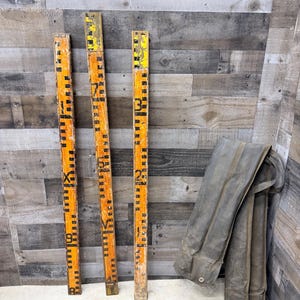 May include: Three vintage orange and black measuring sticks with black numerals and markings. The sticks are leaning against a weathered wood wall. A folded, gray, work jacket hangs to the right.