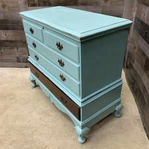 Pu&ograve; includere: Un antico baule di colore azzurro chiaro con quattro cassetti e un coperchio superiore. Il baule ha una base in legno scuro e piedini decorativi.
