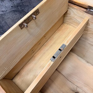 Op de afbeelding: Een open, antieke houten kist met een lichtgekleurde afwerking. Het interieur onthult een kleine lade en metalen hardware. De kist lijkt versleten, met zichtbare houtnerf en tekenen van ouderdom, wat suggereert dat het een vintage item is.