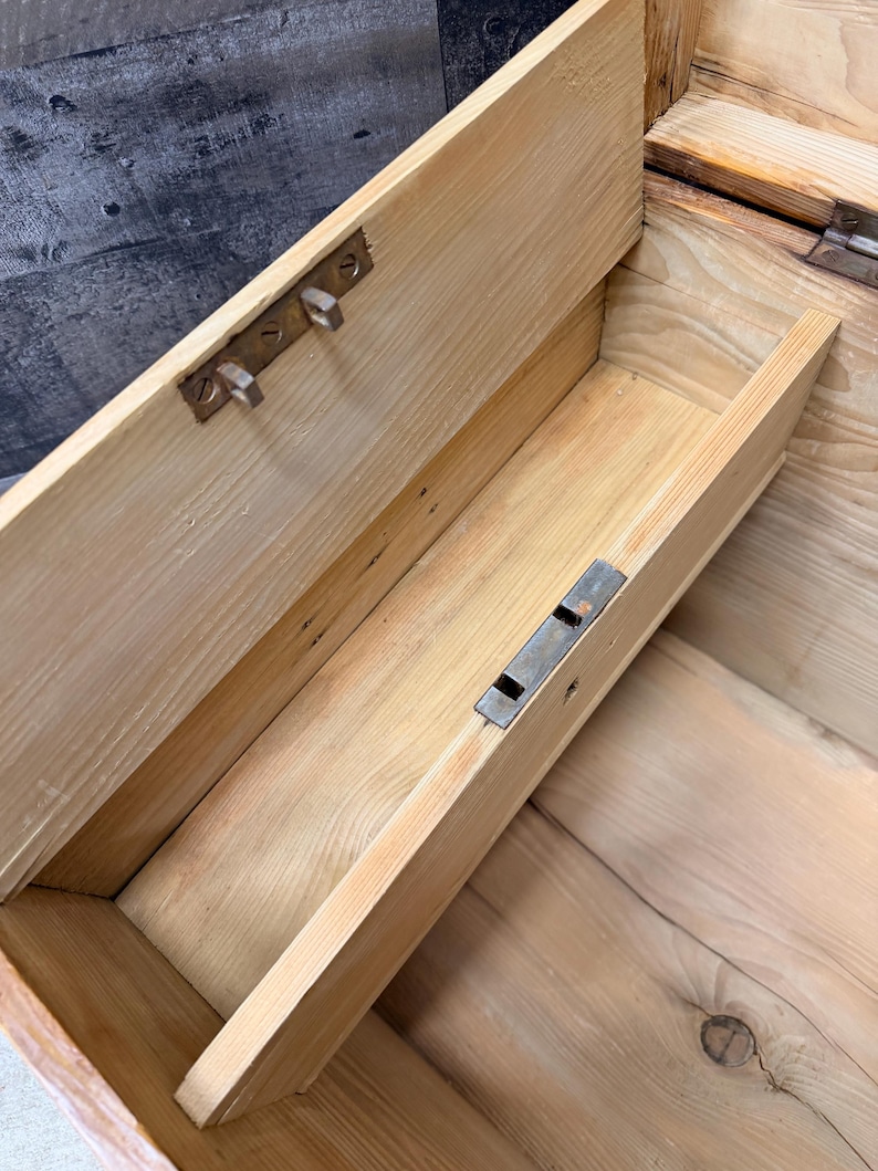 Op de afbeelding: Een open, antieke houten kist met een lichtgekleurde afwerking. Het interieur onthult een kleine lade en metalen hardware. De kist lijkt versleten, met zichtbare houtnerf en tekenen van ouderdom, wat suggereert dat het een vintage item is.