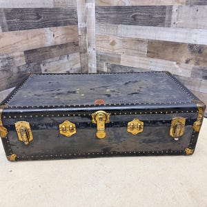 May include: A vintage black trunk with brass hardware. The trunk has a rectangular shape with rounded corners and is adorned with brass locks, hinges, and corner protectors. The trunk's exterior shows signs of wear, adding to its antique charm.
