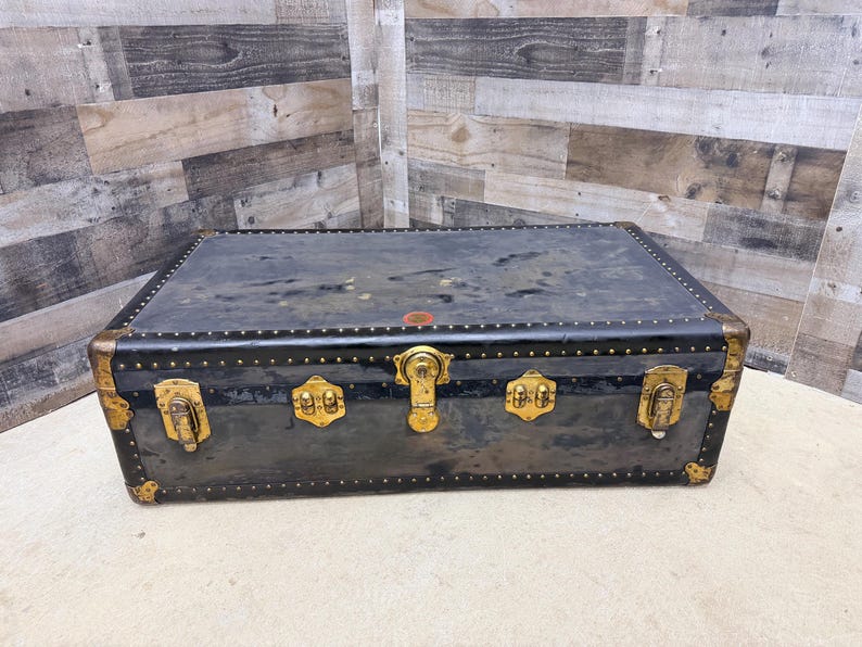 May include: A vintage black trunk with brass hardware. The trunk has a rectangular shape with rounded corners and is adorned with brass locks, hinges, and corner protectors. The trunk's exterior shows signs of wear, adding to its antique charm.