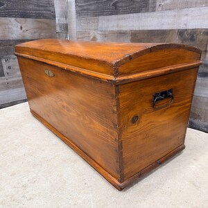 Op de afbeelding: Een houten kist met een gebogen deksel en metalen beslag. De kist is gemaakt van hout met een warme, roodbruine afwerking. Het heeft een rechthoekige vorm met een gebogen bovenkant en een metalen handvat aan de zijkant.