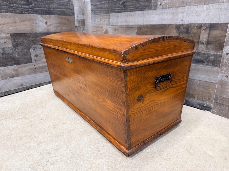 Op de afbeelding: Een houten kist met een gebogen deksel en metalen beslag. De kist is gemaakt van hout met een warme, roodbruine afwerking. Het heeft een rechthoekige vorm met een gebogen bovenkant en een metalen handvat aan de zijkant.