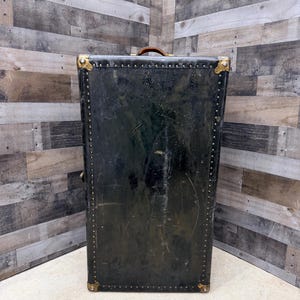 May include: A tall, vintage black trunk with brass corner protectors and rivet detailing. The trunk has a brown leather handle and shows signs of wear, suggesting its age and use. The trunk is a storage item.