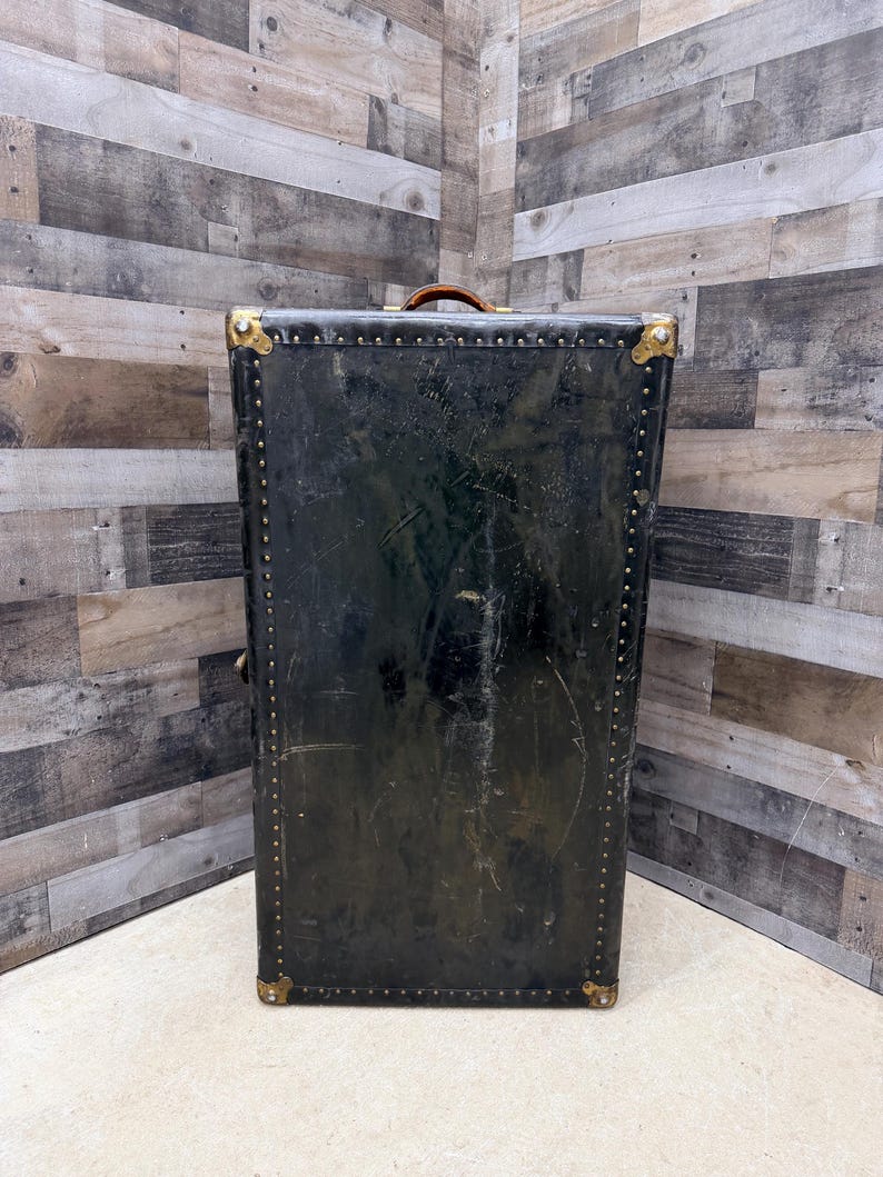 May include: A tall, vintage black trunk with brass corner protectors and rivet detailing. The trunk has a brown leather handle and shows signs of wear, suggesting its age and use. The trunk is a storage item.
