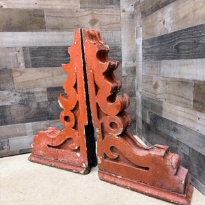 May include: Two antique, ornate wooden architectural brackets. The brackets are painted a weathered orange-red with white accents. Each bracket features a decorative scroll design and is approximately 18 inches tall.