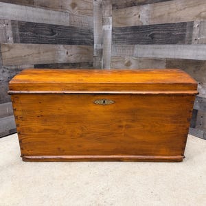 Op de afbeelding: Een houten kist met een gebogen bovenkant en een messing sleutelgatdeksel. De kist is gemaakt van lichtbruin hout met zichtbare nerf en is rechthoekig van vorm. De kist is een opbergcontainer.
