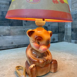 May include: A vintage ceramic bear lamp with a brown and orange finish. The lamp features a lampshade with a red and yellow design. The lamp has a cord and a wooden light socket.