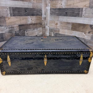 May include: A rectangular black trunk with brass-colored hardware and rivets. The trunk has a slightly distressed appearance, with visible wear and tear. The corners are reinforced with metal, and the trunk has three clasps.