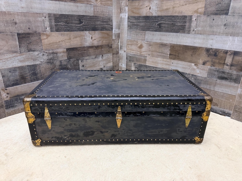 May include: A rectangular black trunk with brass-colored hardware and rivets. The trunk has a slightly distressed appearance, with visible wear and tear. The corners are reinforced with metal, and the trunk has three clasps.