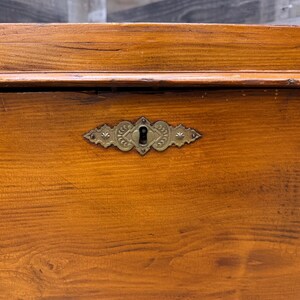 Op de afbeelding: Close-up van een houten kist met een decoratieve messing sleutelgatcover. De kist is gemaakt van warm getint hout met zichtbare nerf. De sleutelgatcover heeft een sierlijk ontwerp met een sleutelgat in het midden.