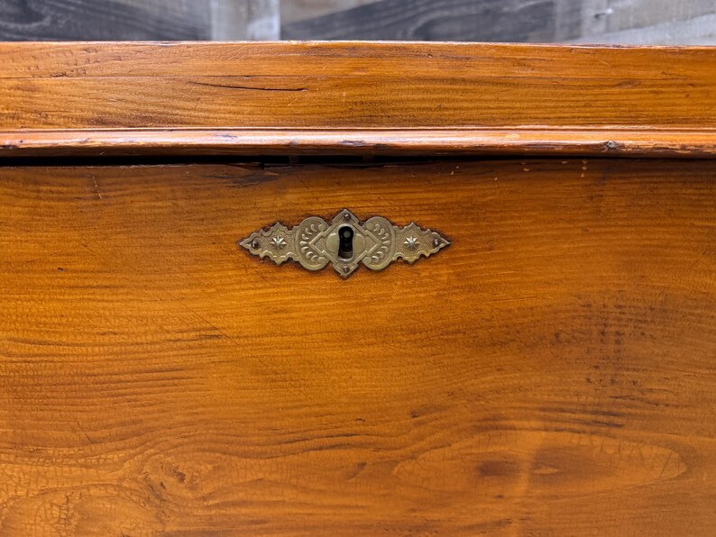 Op de afbeelding: Close-up van een houten kist met een decoratieve messing sleutelgatcover. De kist is gemaakt van warm getint hout met zichtbare nerf. De sleutelgatcover heeft een sierlijk ontwerp met een sleutelgat in het midden.