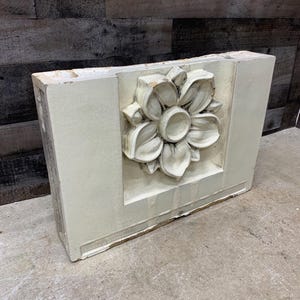 May include: A white plaster architectural element with a raised floral design. The design features a large flower with multiple petals and a center circle. The element is rectangular and has a rough texture.