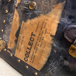May include: Close-up of a vintage black trunk with brass hardware and a worn paper label. The label reads "COLLECT" and "RAILWAY EXPRESS". The trunk's corners are reinforced with metal and secured with rivets, showing signs of age and use.
