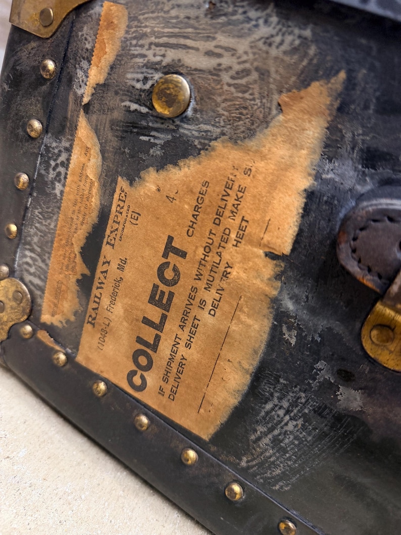 May include: Close-up of a vintage black trunk with brass hardware and a worn paper label. The label reads "COLLECT" and "RAILWAY EXPRESS". The trunk's corners are reinforced with metal and secured with rivets, showing signs of age and use.