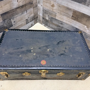 May include: A vintage black trunk with brass hardware and corner protectors. The trunk has a rectangular shape and is adorned with decorative metal accents and a small red emblem. The trunk is a classic piece of luggage.
