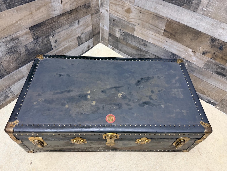 May include: A vintage black trunk with brass hardware and corner protectors. The trunk has a rectangular shape and is adorned with decorative metal accents and a small red emblem. The trunk is a classic piece of luggage.