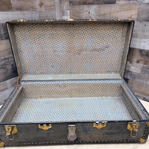 May include: An open, antique trunk with a dark exterior and brass hardware. The interior is lined with a patterned blue and gold fabric. The trunk has a rectangular shape and is designed for storage.