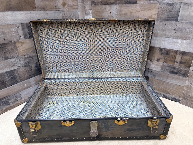 May include: An open, antique trunk with a dark exterior and brass hardware. The interior is lined with a patterned blue and gold fabric. The trunk has a rectangular shape and is designed for storage.