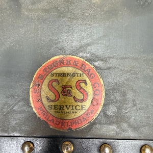 May include: Close-up of a vintage trunk detail. The image shows a circular label with the text "S & S SERVICE" and "STRENGTH & BAG CO." in red and gold. The trunk has brass rivets and a dark gray surface.