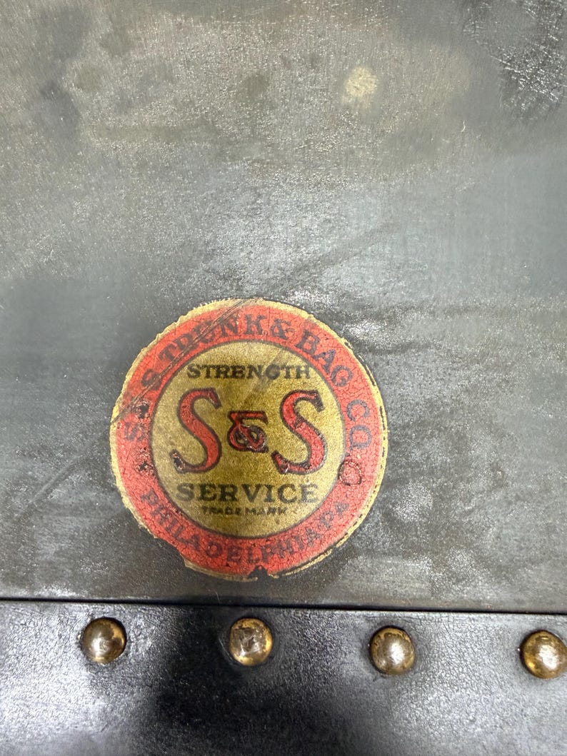 May include: Close-up of a vintage trunk detail. The image shows a circular label with the text "S & S SERVICE" and "STRENGTH & BAG CO." in red and gold. The trunk has brass rivets and a dark gray surface.