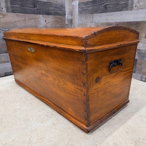 Op de afbeelding: Een grote, antieke houten kist met een gebogen deksel. De kist is gemaakt van warm getint hout met zichtbare nerf en heeft een metalen slot en handvat. De kist is rechthoekig met een licht gebogen bovenkant.
