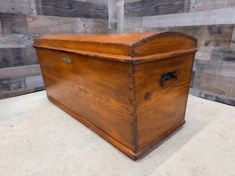 Op de afbeelding: Een grote, antieke houten kist met een gebogen deksel. De kist is gemaakt van warm getint hout met zichtbare nerf en heeft een metalen slot en handvat. De kist is rechthoekig met een licht gebogen bovenkant.