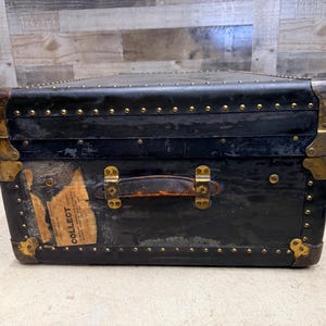May include: A vintage black trunk with brass hardware and a leather handle. The trunk has a rectangular shape with rounded corners and is adorned with small, evenly spaced rivets. A label with the word "COLLECT" is visible on the front.