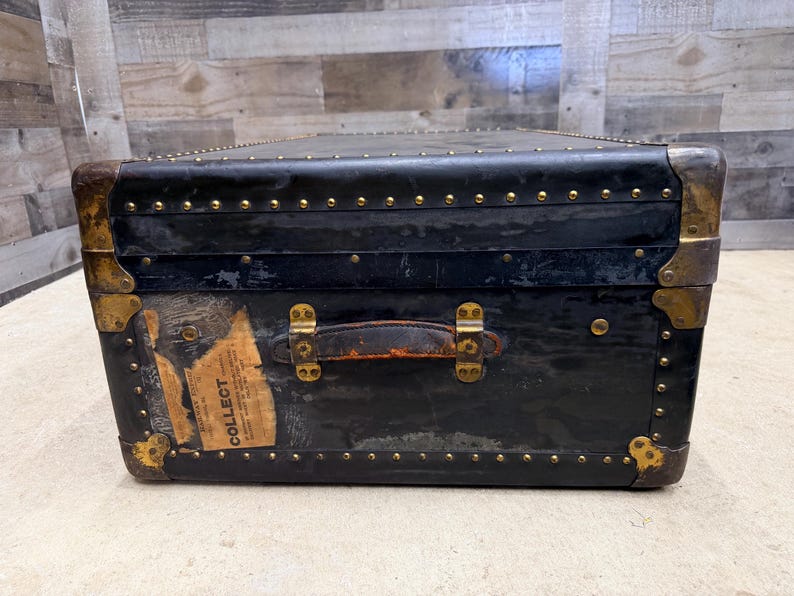 May include: A vintage black trunk with brass hardware and a leather handle. The trunk has a rectangular shape with rounded corners and is adorned with small, evenly spaced rivets. A label with the word "COLLECT" is visible on the front.