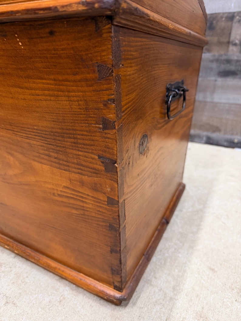 Op de afbeelding: Een houten kist met een gebogen bovenkant en een metalen handvat. De kist is geconstrueerd met zwaluwstaartverbindingen en heeft een warme, roodbruine afwerking. De houtnerf is zichtbaar.