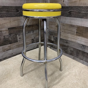 May include: A retro-style bar stool with a bright yellow padded seat and a chrome-plated steel frame. The seat is round with a silver trim. The stool has a footrest and four curved legs. The stool is designed for a kitchen or bar.