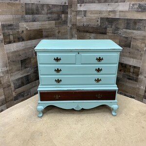 Pu&ograve; includere: Un baule in legno dipinto di azzurro con quattro cassetti e un cassetto inferiore. Il baule ha gambe ornate e ferramenta in ottone.