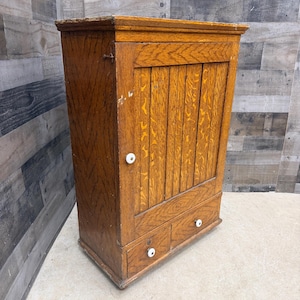 May include: A medium-brown oak wall cabinet featuring a single vertical-slatted door with a white ceramic knob. Below the door are two small drawers, each equipped with a matching white ceramic knob. The wood grain is visible throughout the piece.