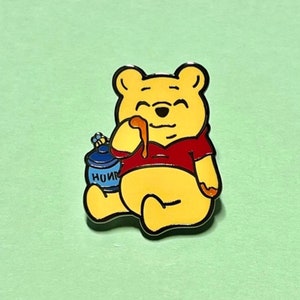 May include: A yellow cartoon bear wearing a red shirt is sitting with a blue honey pot. The bear is licking honey from a dripping honey stick. The honey pot has the word "HONEY" written on it.