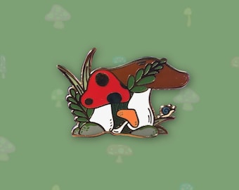 May include: A gold-tone enamel pin featuring a cartoon mushroom with a red cap and white stem, surrounded by green leaves and a small snail. The mushroom is sitting on a brown rock.
