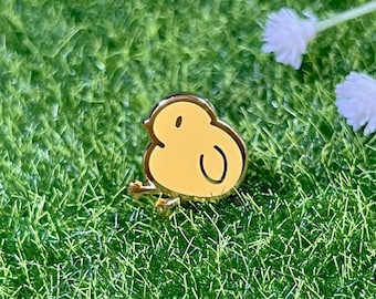 May include: A yellow enamel pin shaped like a cartoon chick with a black outline. The pin is on a green grass background.