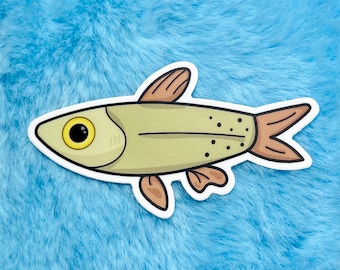 May include: A sticker featuring a cartoon fish with a yellow body, brown fins, and a white outline. The fish has a large, yellow eye and small black dots along its side. The sticker is placed on a blue, fluffy surface.
