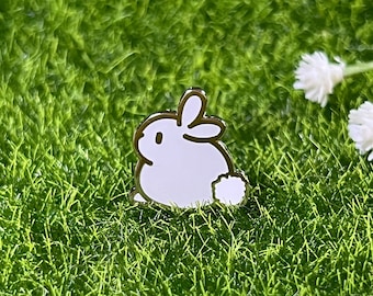May include: A white enamel pin of a cartoon bunny rabbit with a fluffy tail. The pin is on a green grass background.