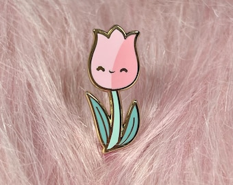 May include: A gold-colored enamel pin featuring a pink and white tulip with a smiling face. The tulip has a green stem and leaves.