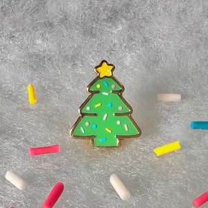 May include: A green enamel Christmas tree pin with a gold star on top and white and yellow sprinkles.