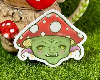 May include: A sticker featuring a green goblin-like creature wearing a red and white mushroom cap. The creature has large eyes, a wide mouth, and a pointed nose. The sticker is on a green grassy surface.
