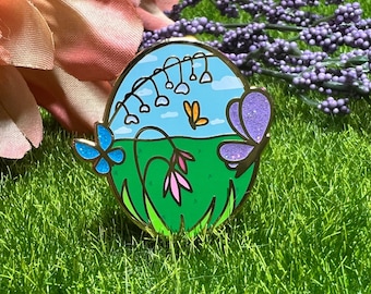May include: An enamel pin with a gold border, featuring a whimsical garden scene. The design includes a blue sky, green grass, and colorful butterflies. The pin showcases a blue butterfly with glitter and a purple butterfly with glitter.