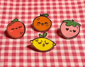 May include: Four enamel pins featuring cartoon fruit with smiling faces. The pins are orange, red, yellow, and pink. The orange pin is a citrus fruit with a green stem. The red pin is an apple with a green stem. The yellow pin is a lemon with a green stem. The pink pin is a strawberry with green leaves.